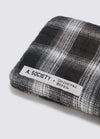 Close-up of A. SOCIETY x Universal Works Olive/Green Kowloon Eyewear case. Plaid pattern in olive and green hues.  A white label with the brand names is stitched on. Shot on a white surface.