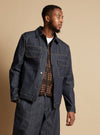 A model poses in an Indigo 13oz Selvedge Denim Trucker Jacket. The dark wash denim jacket is styled with a brown vest. Studio shot on a neutral background.
