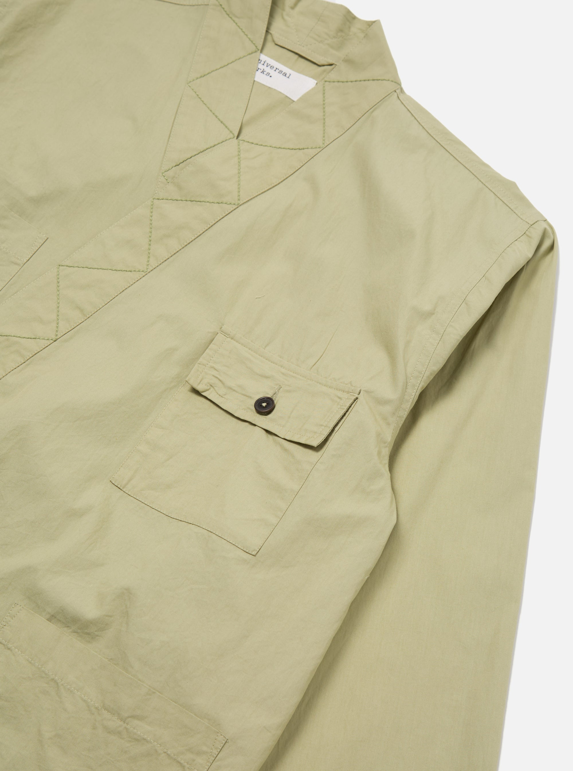 Close-up of the pale green Broadcloth Osaka Work Jacket on a white surface. The broadcloth material has a pocket with a button. The jacket has a unique collar.