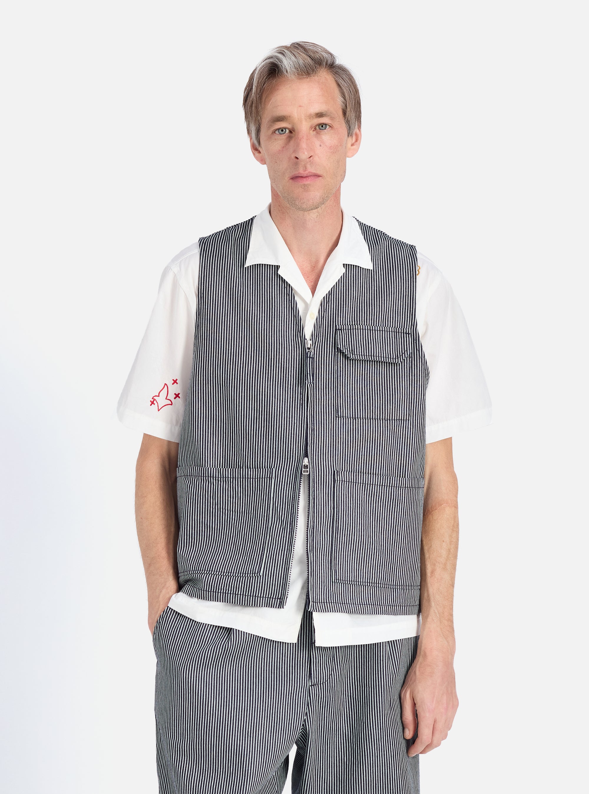 A model on a white background wears the Indigo Hickory Stripe Denim Marine Gilet, a striped vest, over a white shirt. The vest has a zip and pockets. Part of matching trousers are visible.