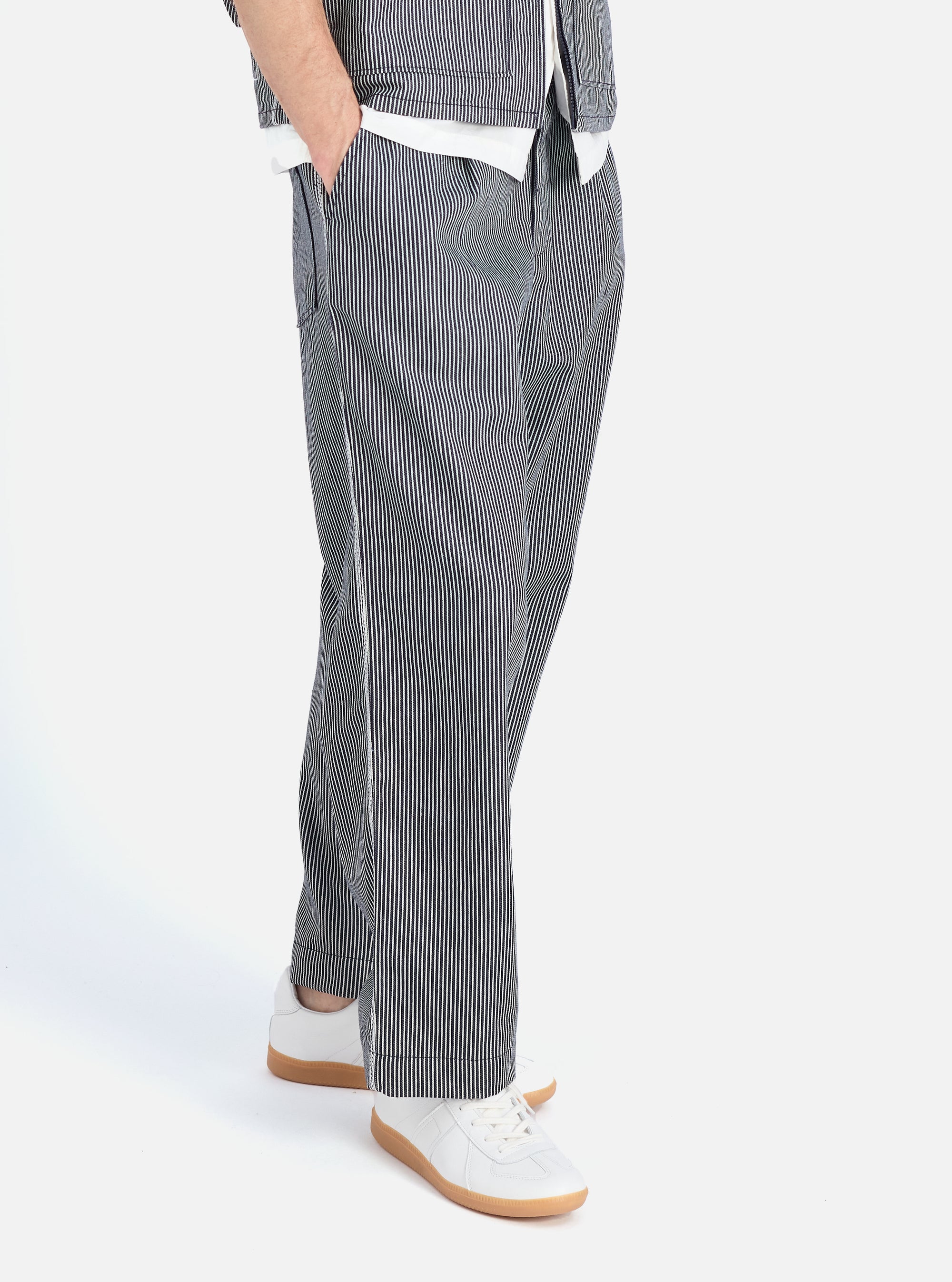 A person wears the Indigo Hickory Stripe Denim Duke Pant in a studio shot. The pants are blue and white striped denim. The background is a bright white.