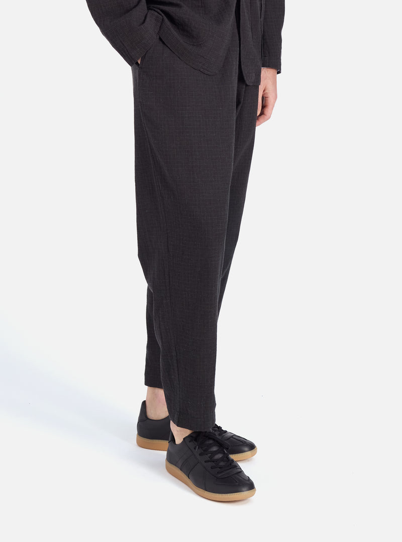 A model on a white background showcases the Charcoal Fine Linen Mix Pleated Track Pant. The pants have a relaxed fit and are paired with black sneakers.