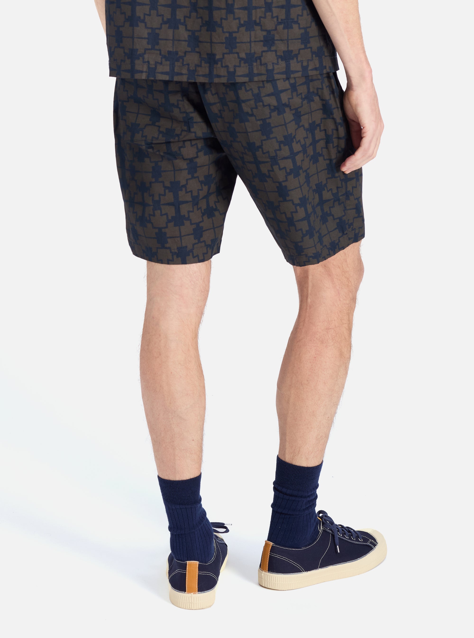 A man stands against a white backdrop, showcasing the Navy Mitla Print Cotton Simple Short. The shorts feature a navy and brown pattern. He wears navy socks and matching sneakers.