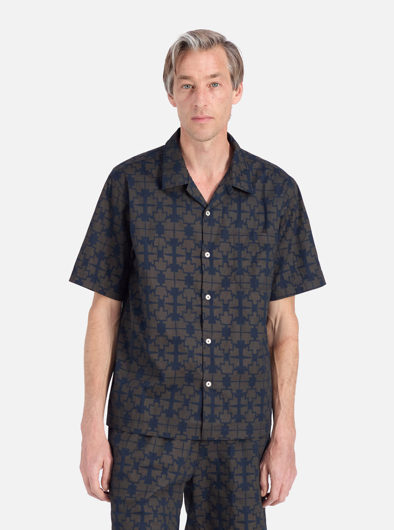 A model stands on a white background wearing the Navy Mitla Print Cotton Road Shirt. The shirt is navy and brown with a geometric print and has short sleeves.
