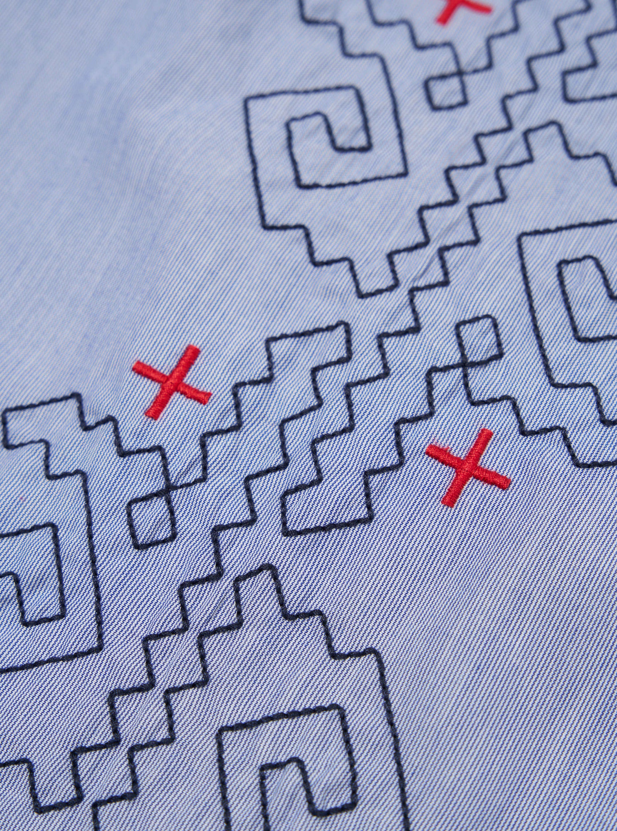 Close-up of the Blue Classic Stripes Road Trip Shirt. Light blue fabric with embroidered black geometric shapes and red crosses. Casual style.