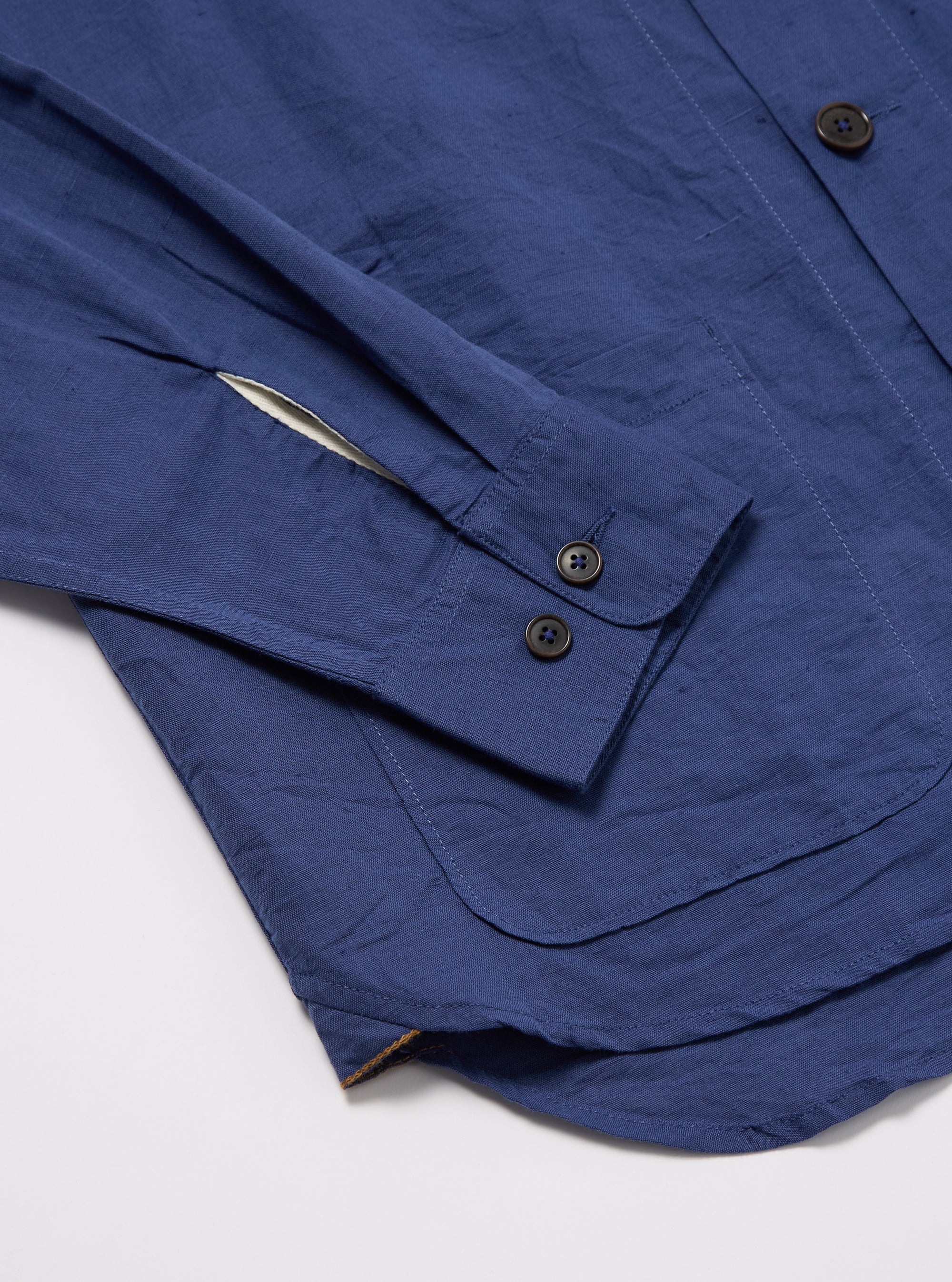 Close-up of the Blue Linen Cotton Shirting Travail Overshirt on a white surface. Shows the blue linen cotton fabric, button details, and pocket stitching.