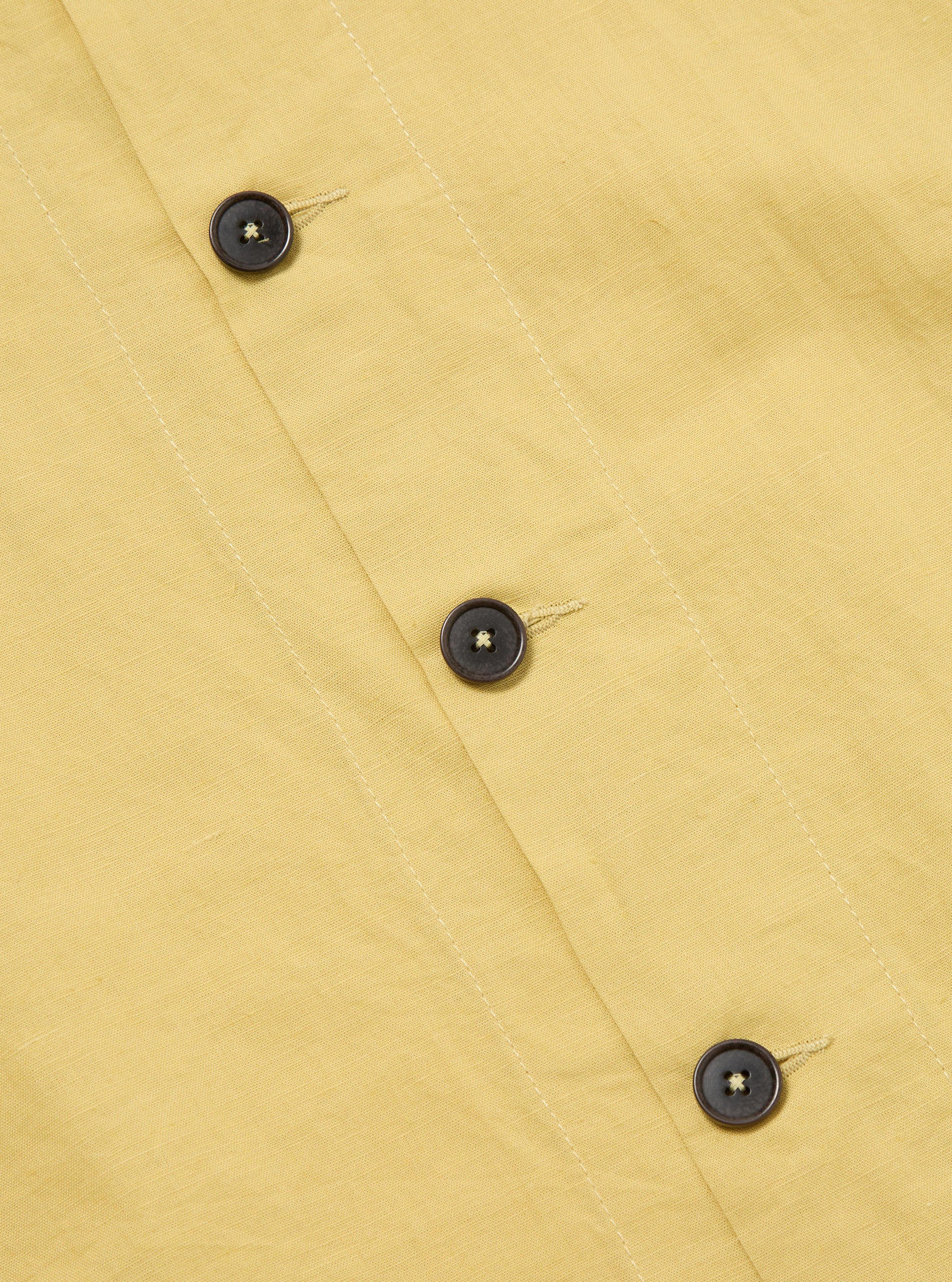 Close-up of the Lemon Linen Cotton Shirting Travail Overshirt, showcasing its yellow linen-cotton blend fabric and dark brown button details on a flat surface.
