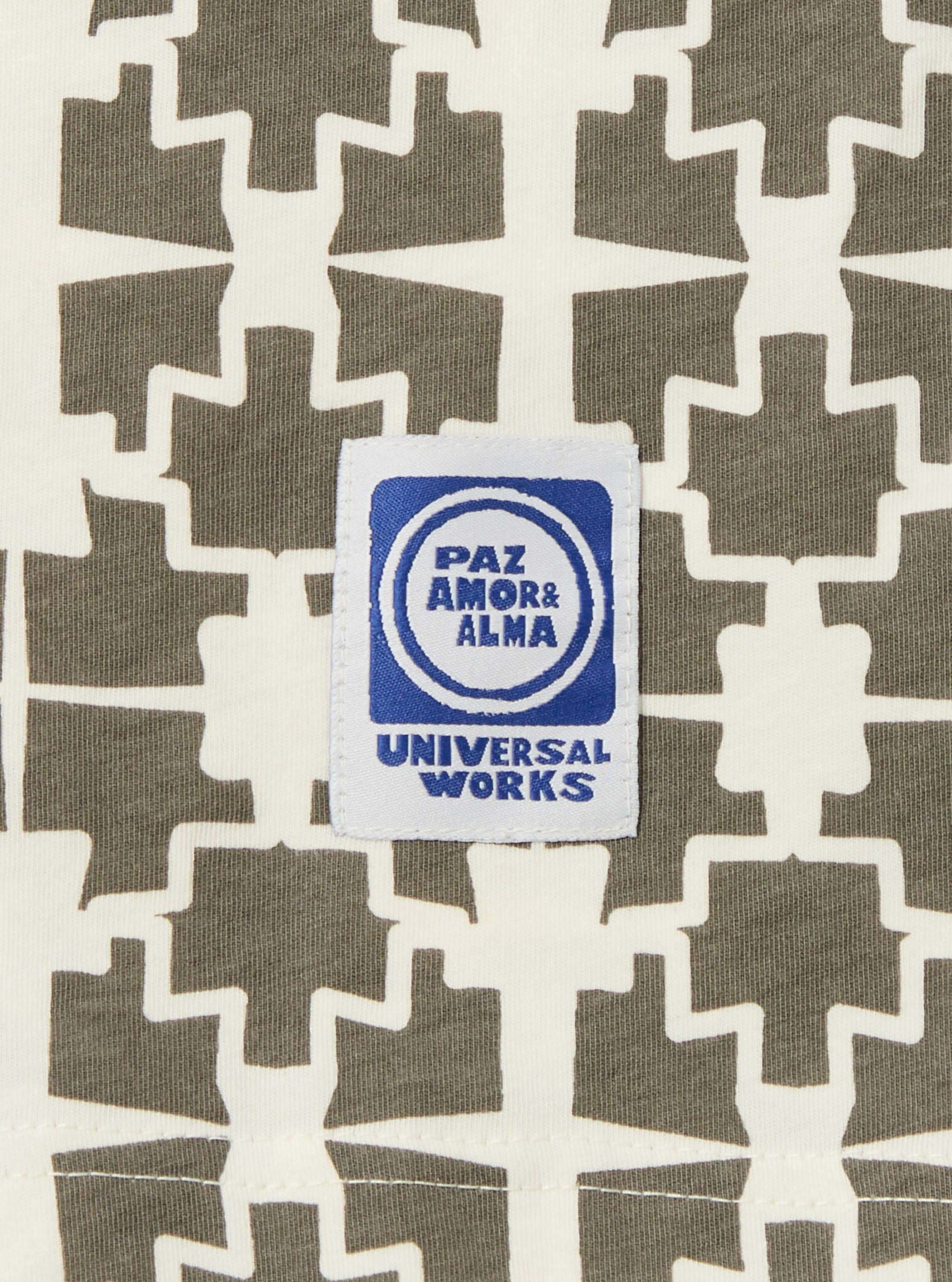 Close-up of the Ecru/Mud Mitla Print Jersey Newlyn Polo, showcasing the geometric pattern and fabric texture. A Universal Works label is visible. 