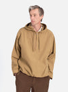 A man poses in the Dark Sand Peach Recycled Nylon Liam Jacket. The jacket is a relaxed fit, with a hood and half-zip. He wears brown pants. Shot on a white background.