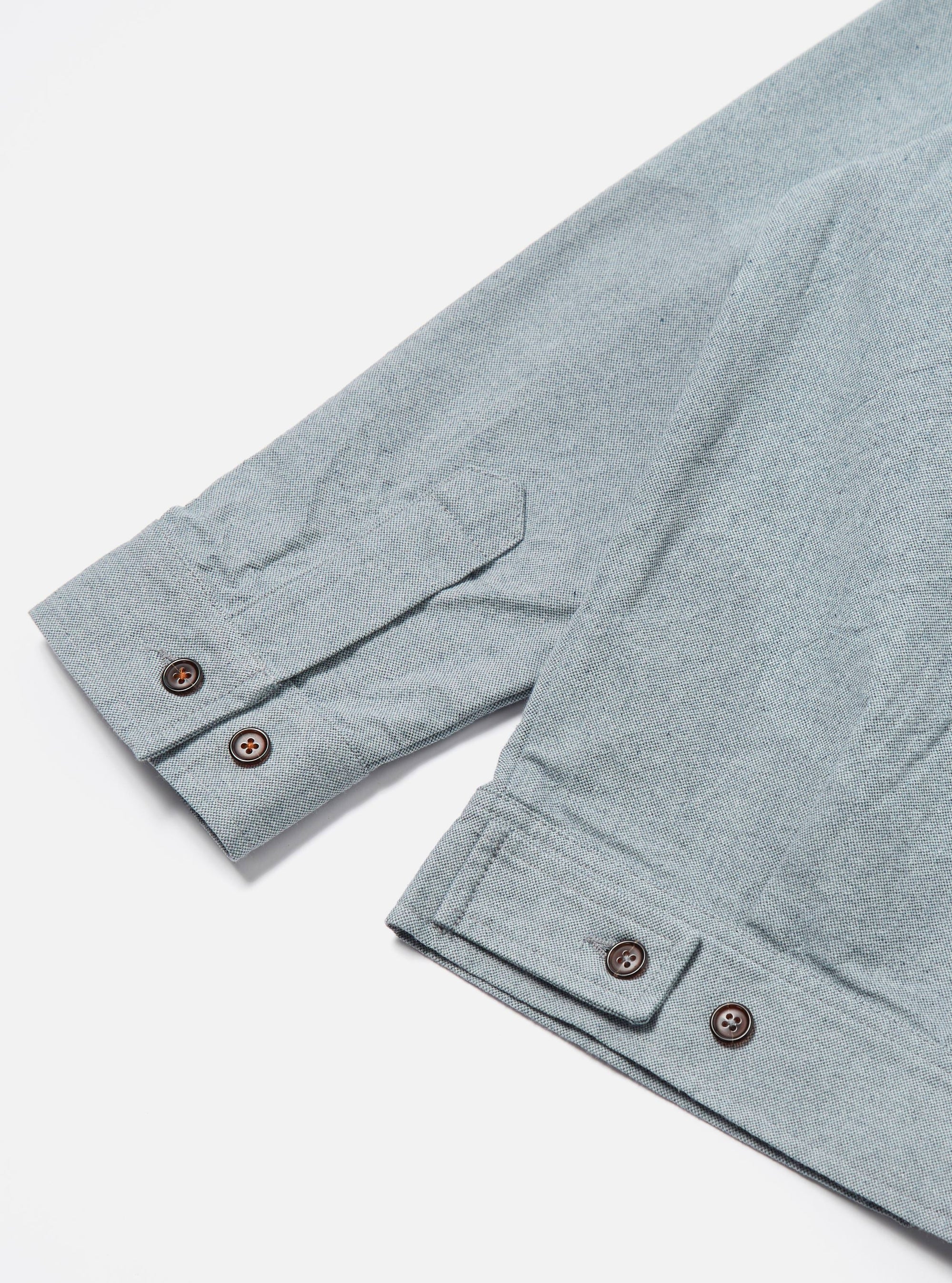 Close-up of the Indigo Reworked Jean Cloth Rose Bowl Jacket sleeve on a white surface. Light blue denim with brown buttons.