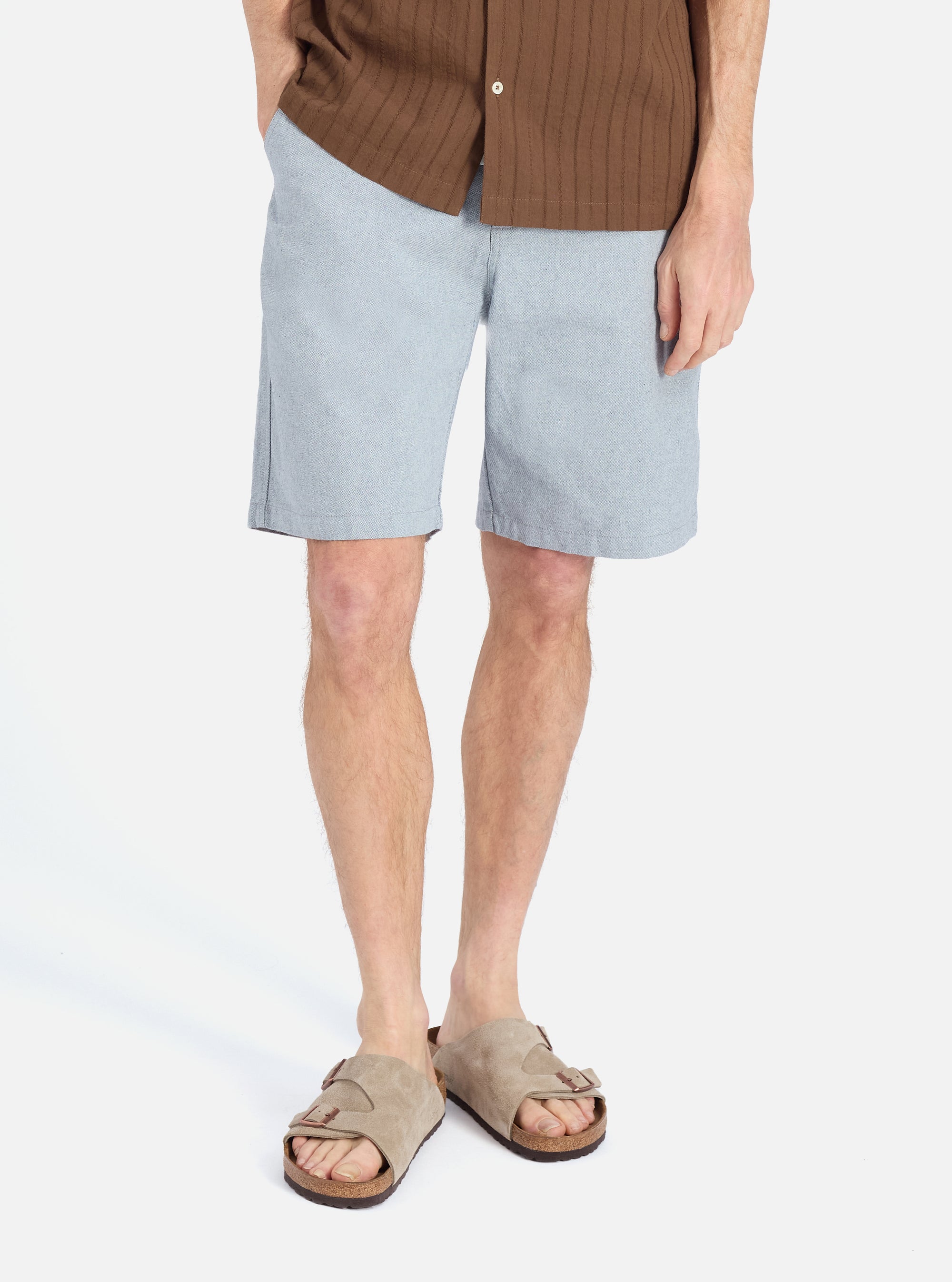 A model on a white background showcases the Indigo Reworked Jean Cloth Alva Short. The light blue shorts are paired with a brown shirt and sandals for a casual look.