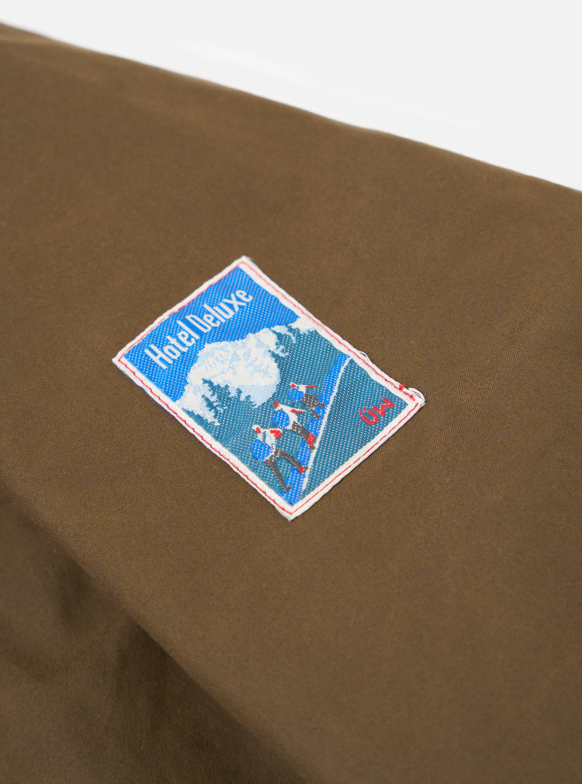 Close up of the brown British Wax Cotton Boston Parka showing the fabric and a Hotel Deluxe patch on a white background.