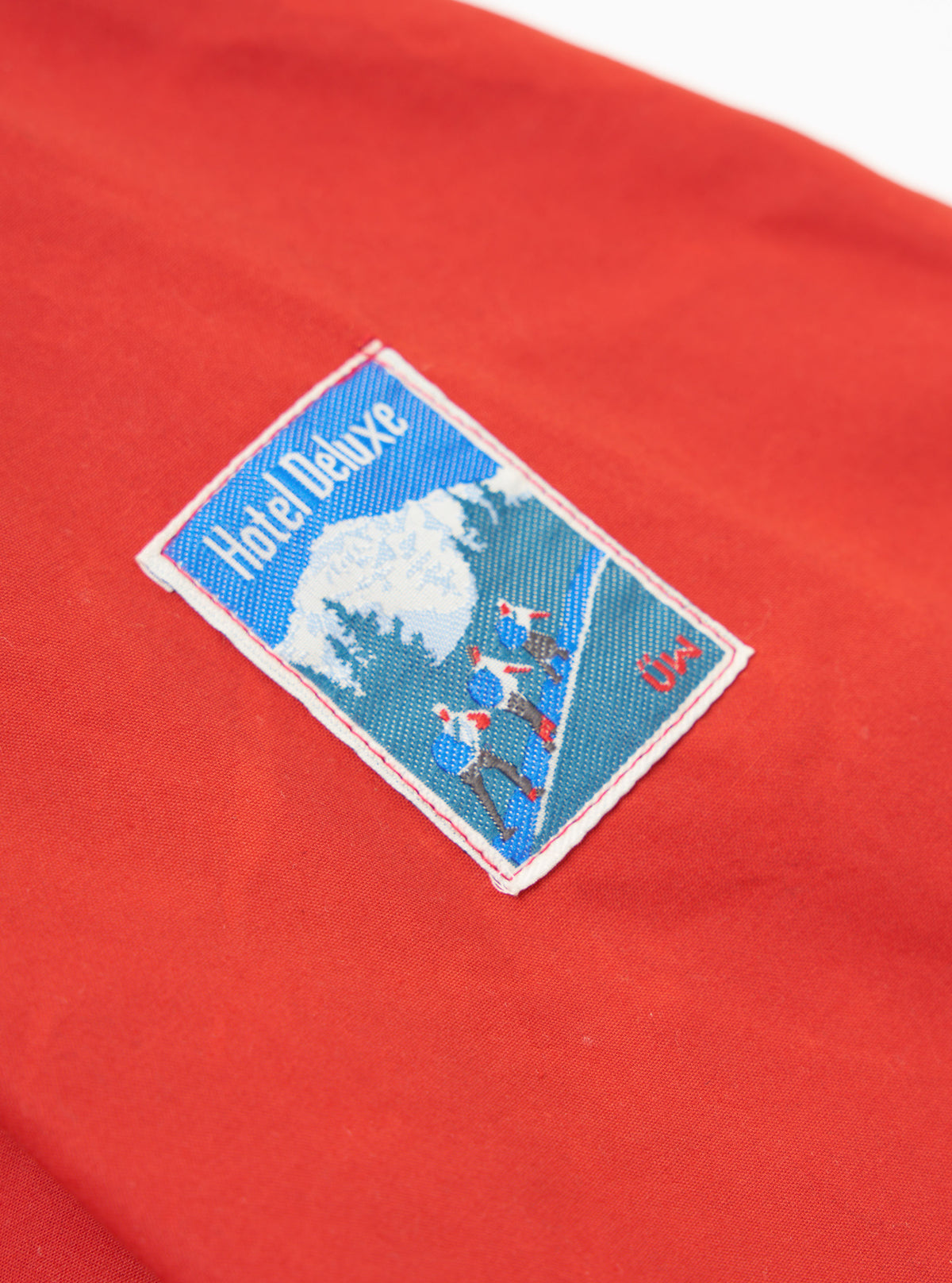 Close-up of the orange British Wax Cotton Boston Parka showing the 'Hotel Deluxe' patch on the sleeve. The patch is blue and white with a red border.