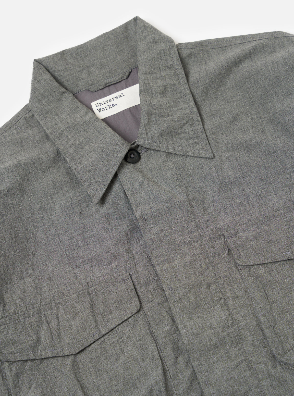 Close-up of the Charcoal Organic Chambray Parachute Field Jacket on a white surface. The jacket is charcoal gray and made of chambray fabric. It features a button closure and two front pockets.