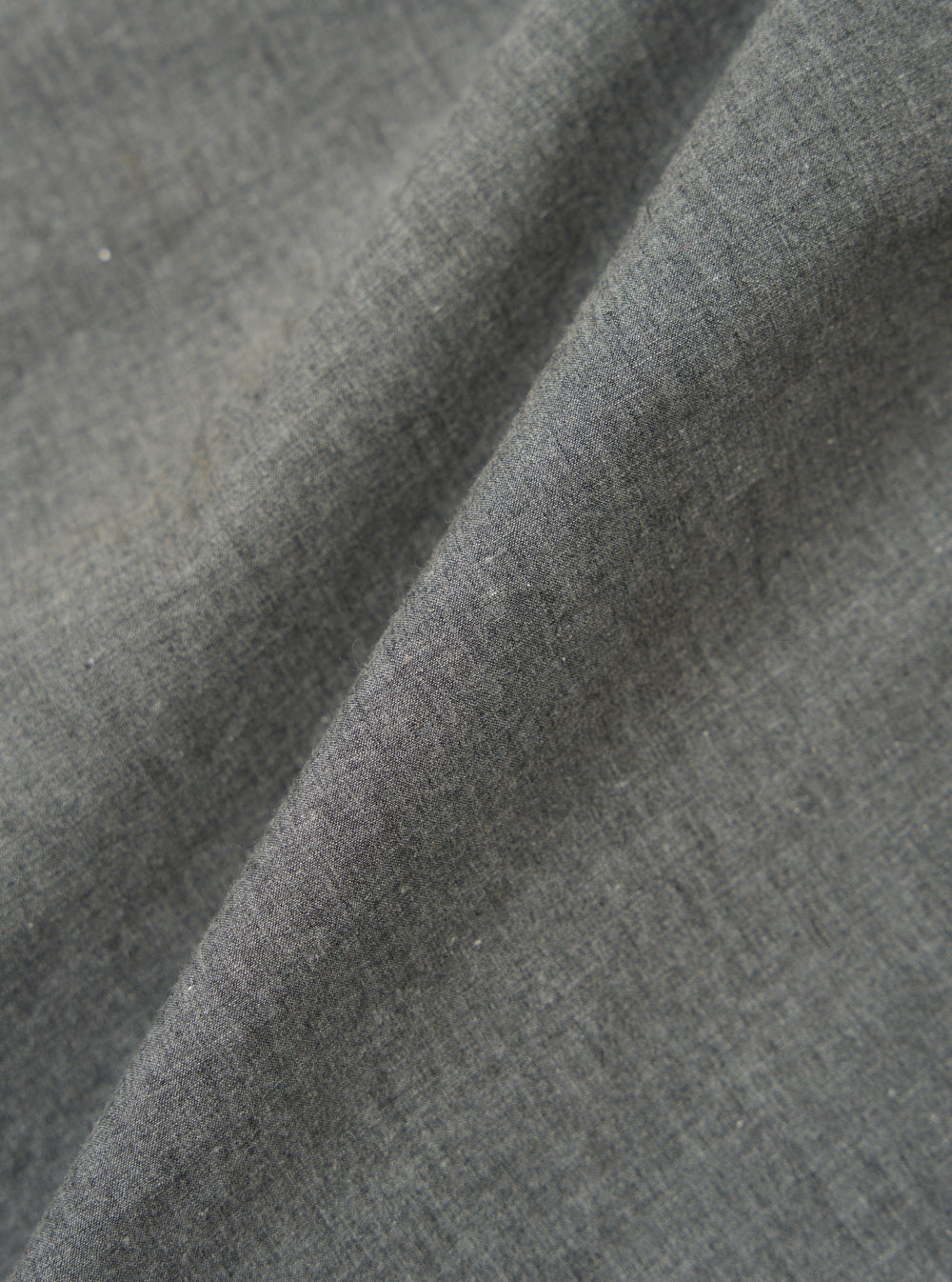 Close-up of the Charcoal Organic Chambray Parachute Field Jacket fabric. The charcoal-colored material is folded, showing its texture. The background is a plain surface.
