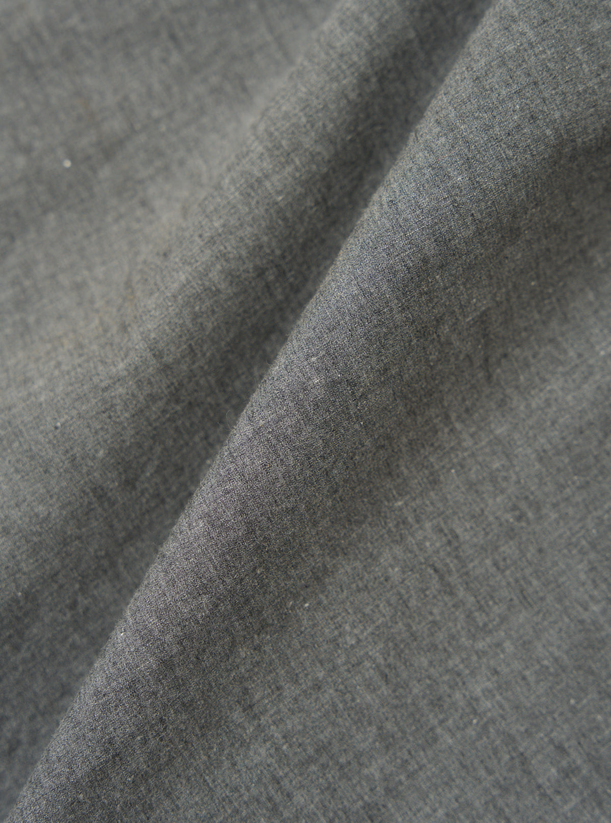 Close-up of the Charcoal Organic Chambray Parachute Field Jacket fabric. The charcoal-colored material is folded, showing its texture. The background is a plain surface.