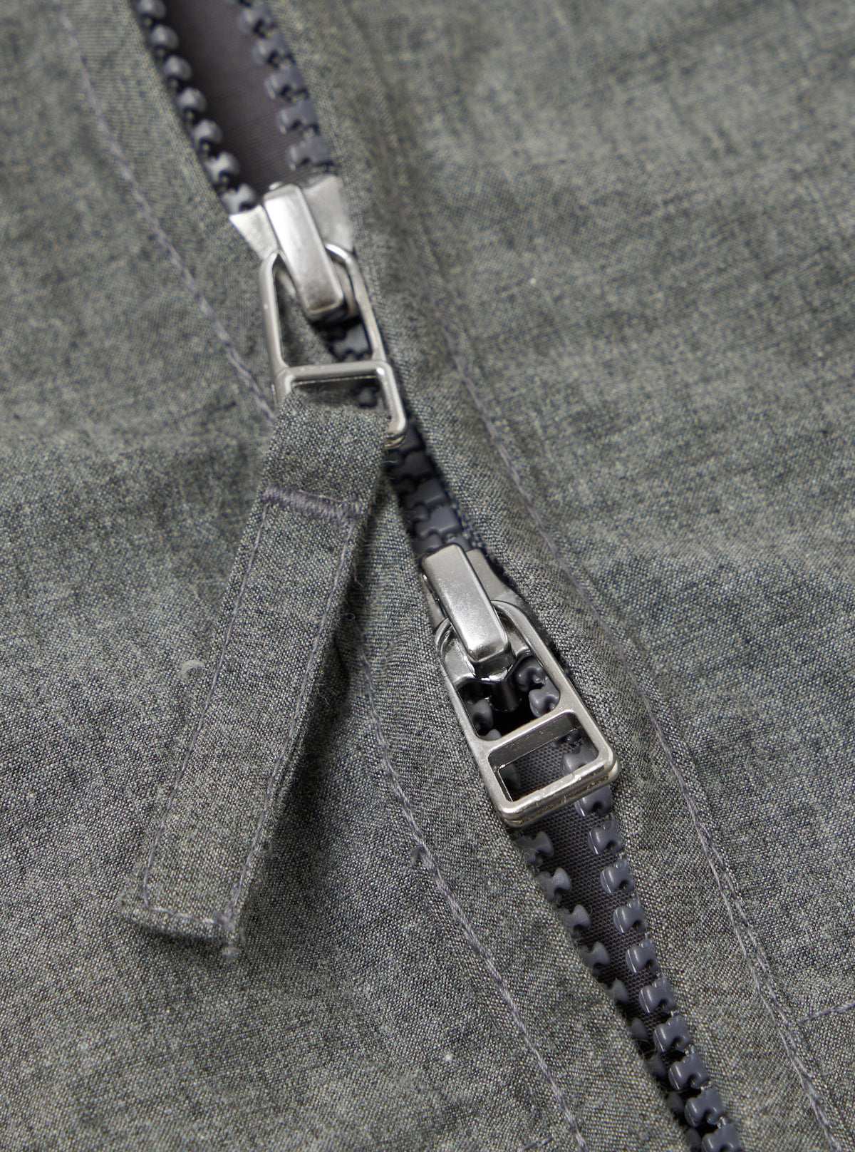 Close-up of the Charcoal Organic Chambray Parachute Liner Jacket. The image shows the zipper with a silver pull tab on the charcoal chambray fabric. 