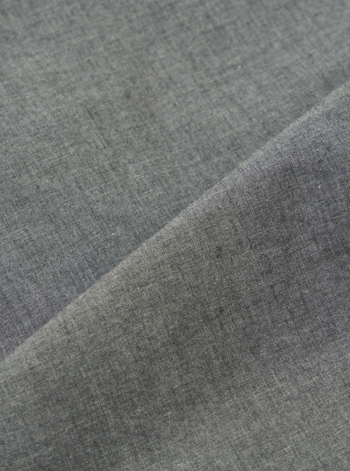 Close-up of the Charcoal Organic Chambray Parachute Liner Jacket fabric. The charcoal material is folded, showing the texture. The background is a plain white surface.