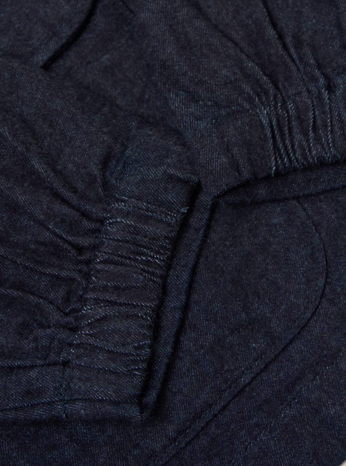 Close-up of the Indigo Brushed Denim Labour Jacket showing the dark blue denim material and pocket detail. The jacket is laid flat on a neutral surface.