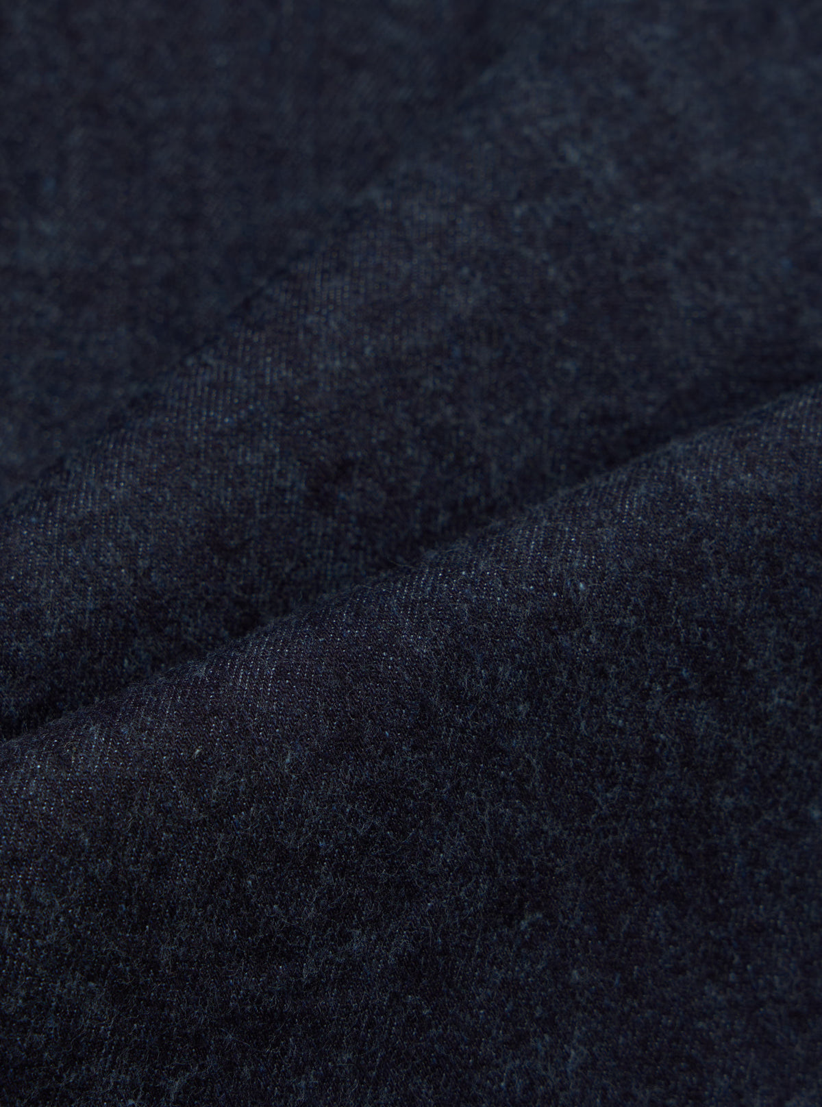 Close-up of the Indigo Brushed Denim Oxford Pant fabric. The indigo denim has a soft, brushed texture. Shot on a plain surface to highlight the material.