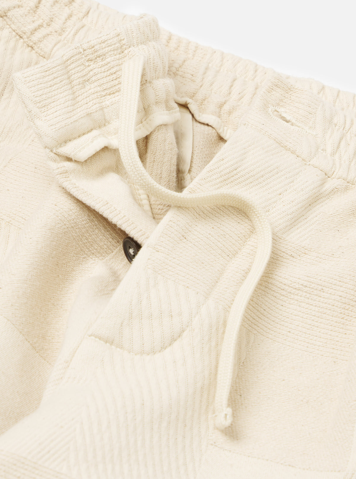 Close-up of the Ecru Patchwork Weave Cotton Pleated Track Pant on a white background. The ecru cotton has a textured, patchwork weave. Drawstring and button details are visible.