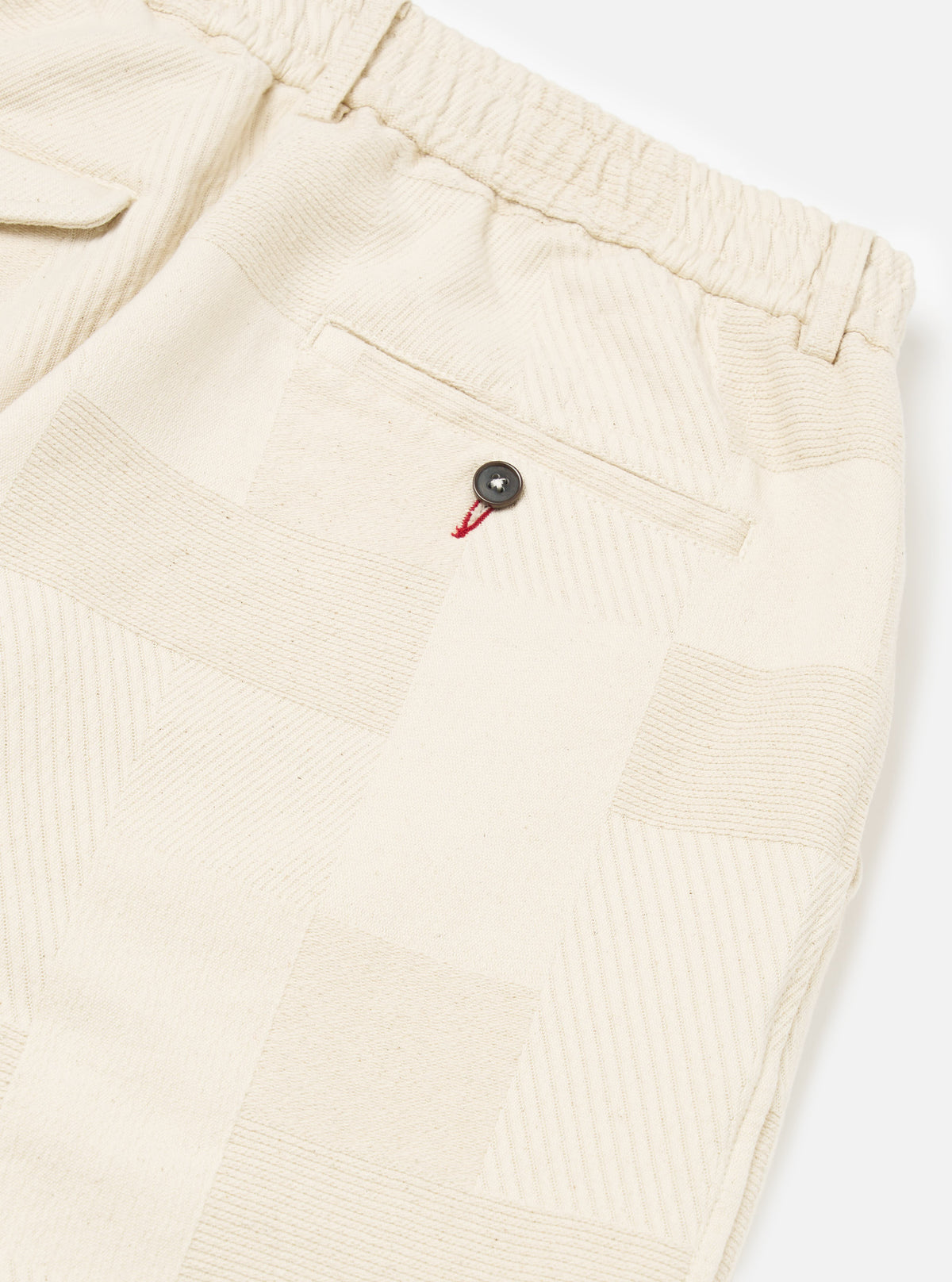 Close-up of the Ecru Patchwork Weave Cotton Pleated Track Pant on a white background. The pants feature a patchwork design and a buttoned pocket.