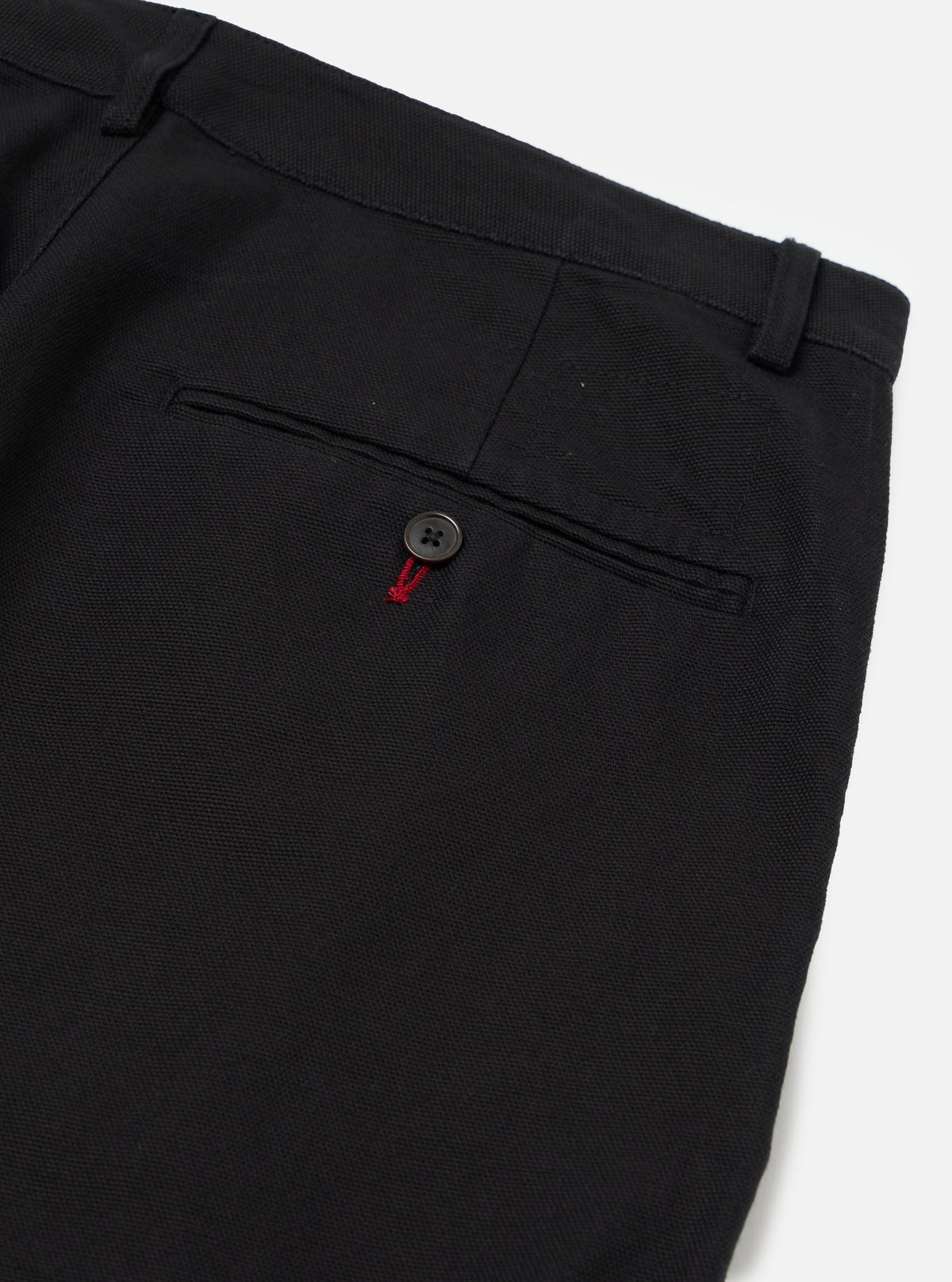 Close-up of the Black Organic Canvas Super Chino showing the back pocket with button detail. The pants are on a white surface.