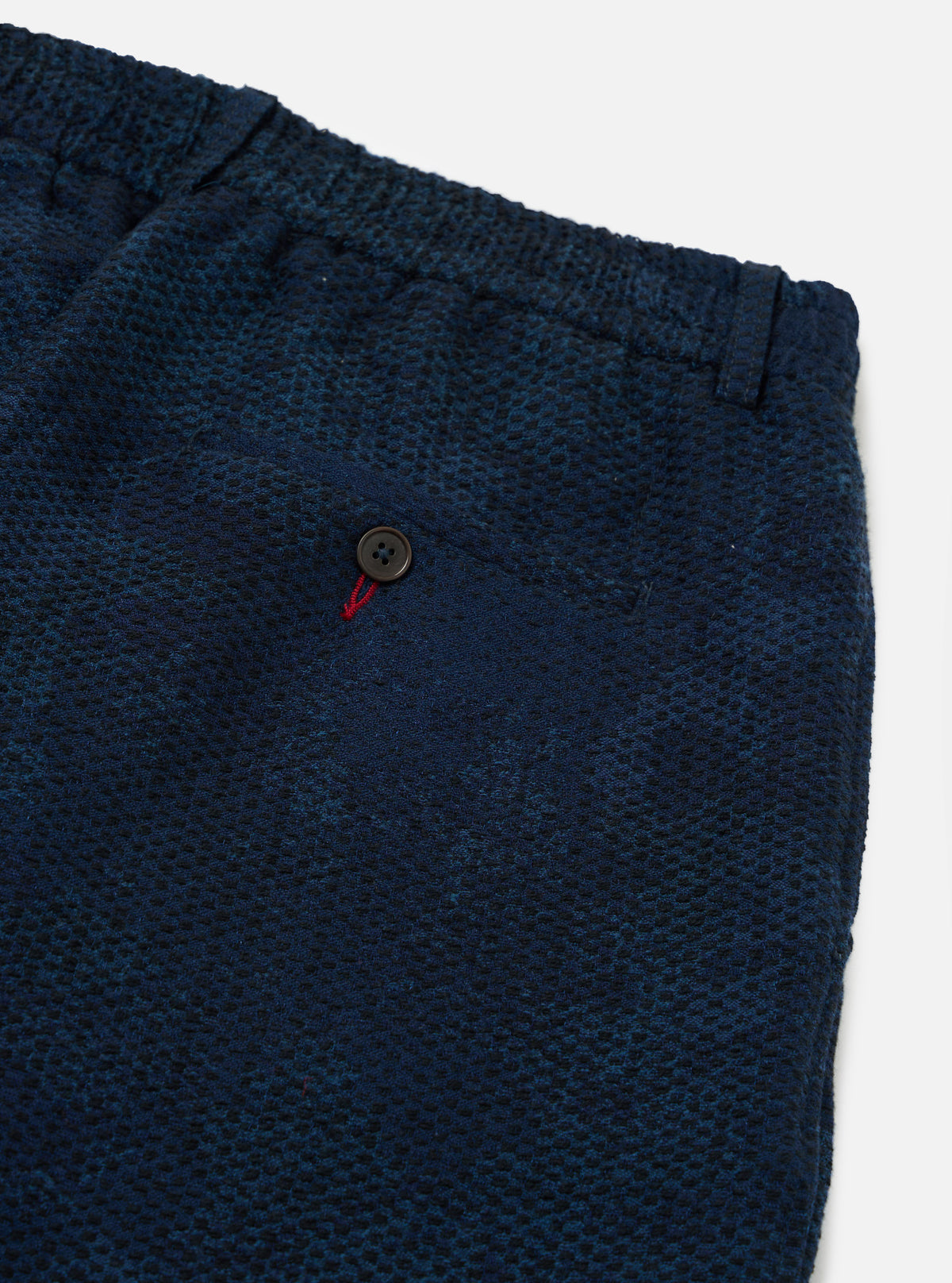 Close-up of Black/Indigo Denim Camo Pleated Track Pant on a white surface. The denim has a subtle camo pattern. Features a buttoned pocket with red stitching detail.