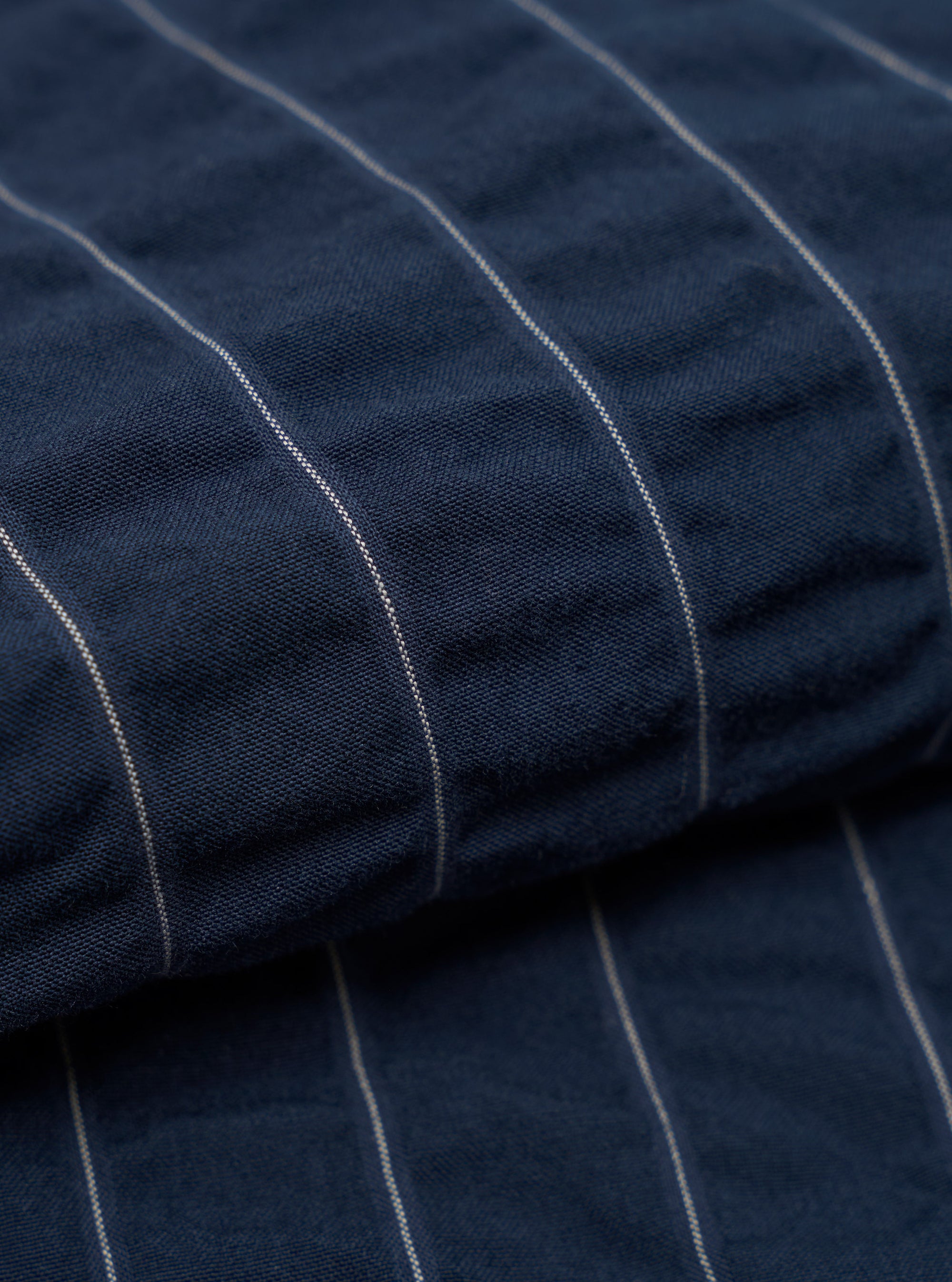 A detailed, close-up fabric swatch of the Navy Chester Stripe Pleated Track Short reveals a pattern of thin, vertical white stripes on a dark navy blue cotton base. The image focuses on the fine texture and weave of the material.