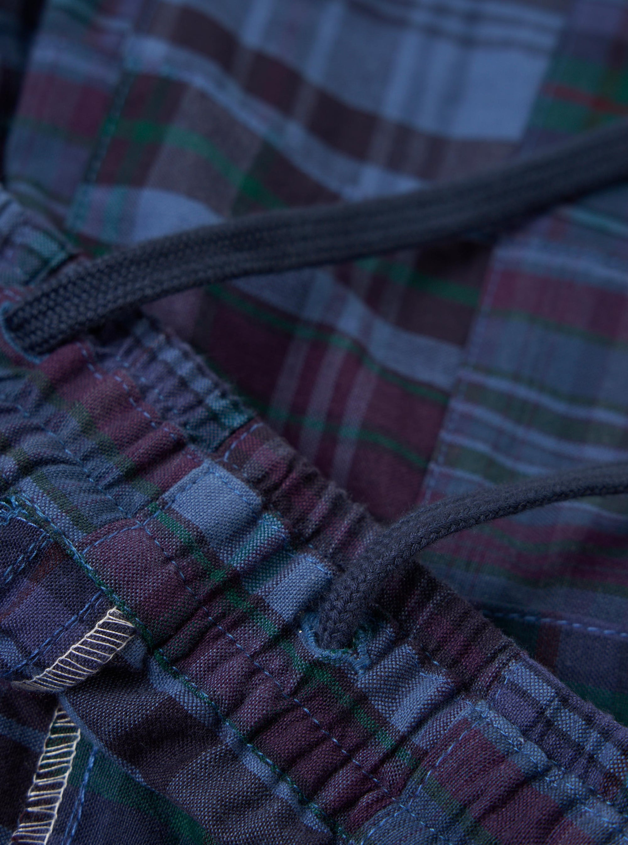 A pair of navy madras patchwork vacation shorts is displayed flat against a plain white background. The shorts feature a patchwork design of various blue and navy plaid fabrics, an elasticated waistband, and a rear patch pocket.