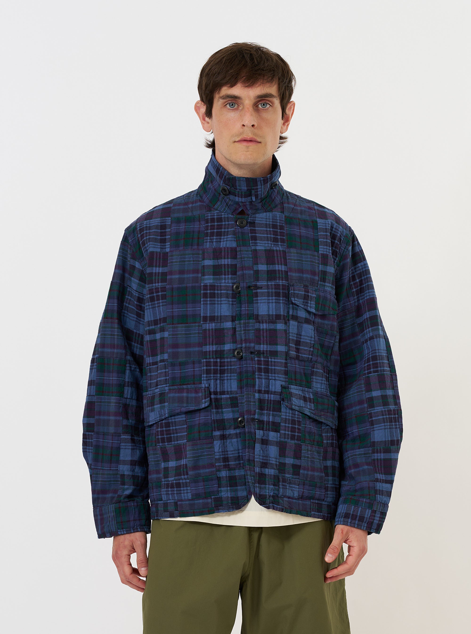 A model is wearing the Navy Madras Patchwork Duty Jacket against a plain white background. The jacket features a vibrant blue and navy madras patchwork pattern, a classic collar, and a full button-down front. The image captures the garment from the waist up, highlighting the detailed patchwork construction and utility-style design.