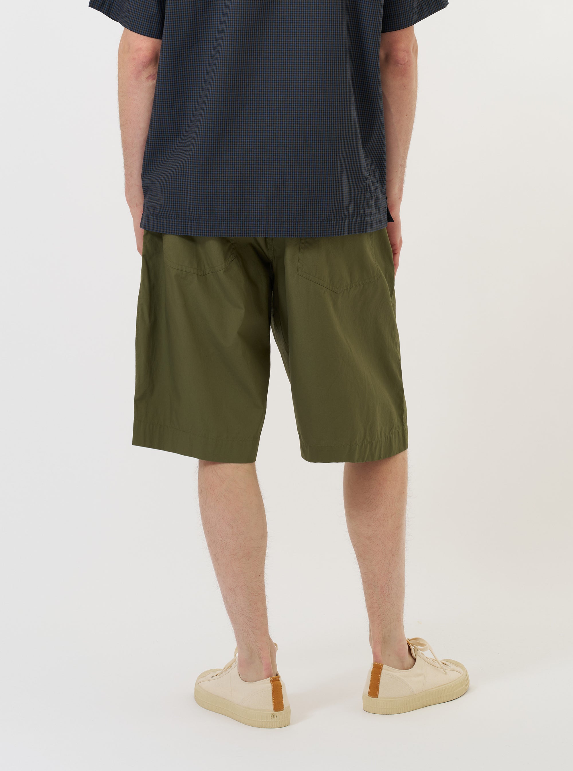 A model is wearing the Olive Broadcloth Vacation Short against a plain white background. The image shows the shorts from the side and rear, highlighting the relaxed fit, mid-thigh length, and a single rear patch pocket. The model is also wearing dark shoes and white socks.