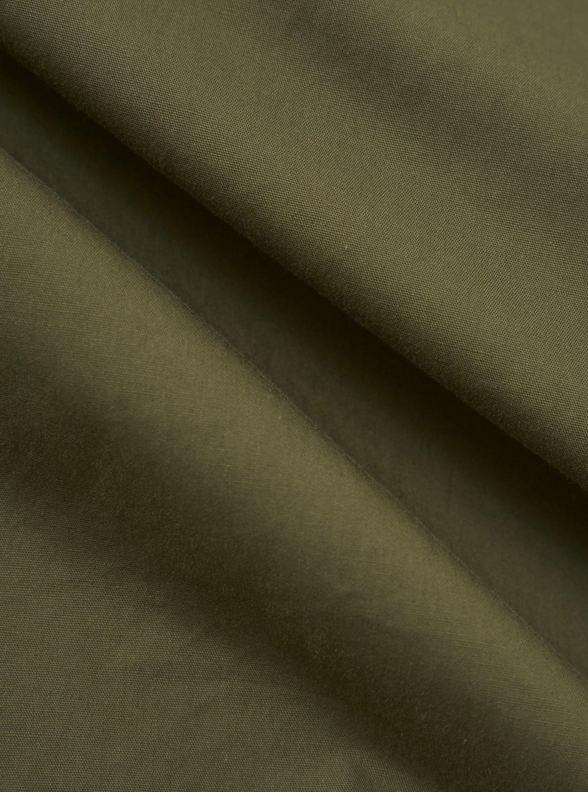 A close-up fabric swatch of the Olive Broadcloth Vacation Short displays the smooth, tight weave of the olive green cotton broadcloth. The image focuses on the uniform color and texture of the material.
