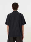 A model is wearing the Navy Broadcloth Camp Shirt against a plain white background. The navy blue short-sleeve shirt is shown from a three-quarter rear perspective, highlighting the smooth broadcloth fabric, the straight hem, and the relaxed fit of the garment as it drapes on the model.