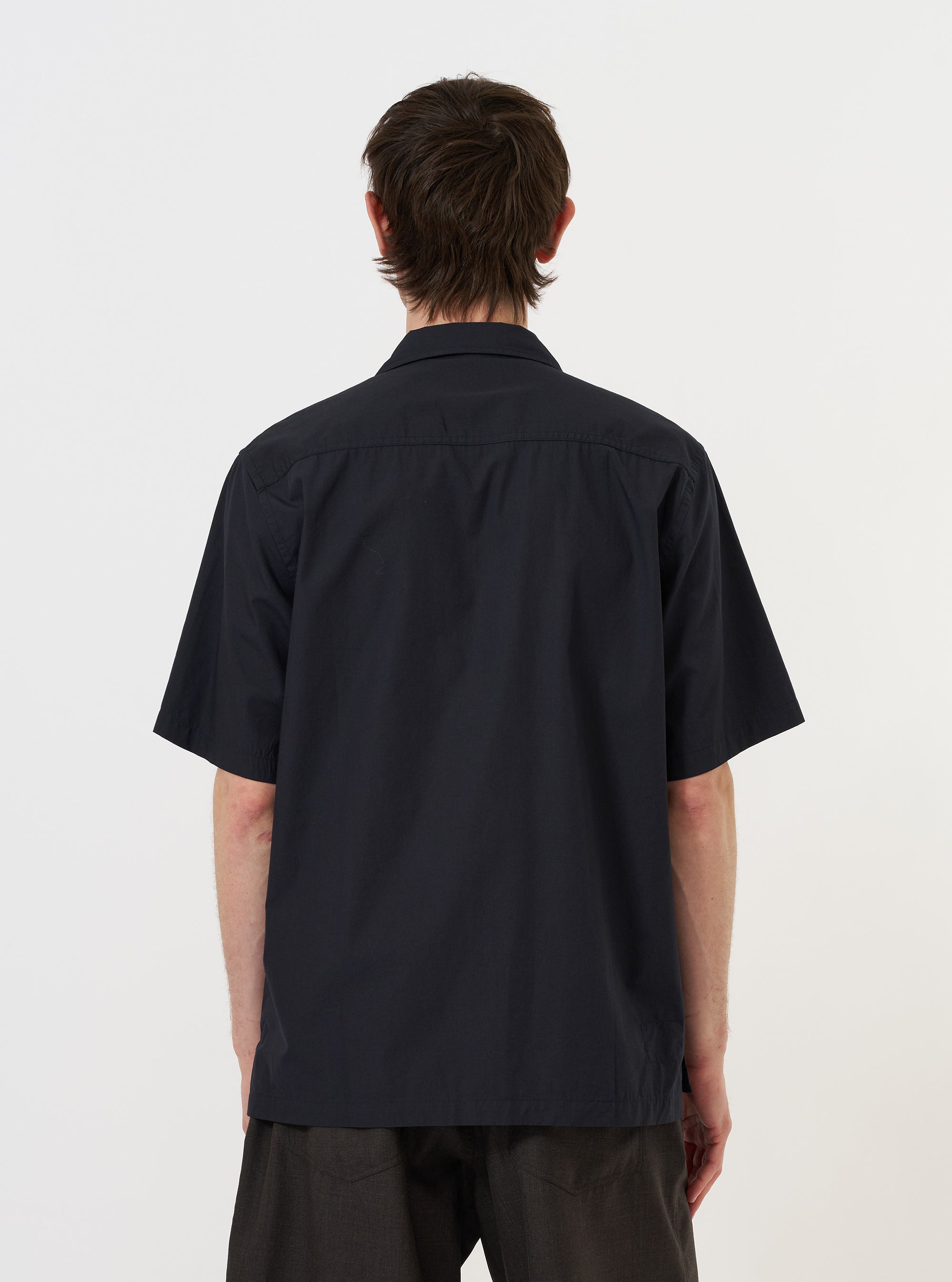 A model is wearing the Navy Broadcloth Camp Shirt against a plain white background. The navy blue short-sleeve shirt is shown from a three-quarter rear perspective, highlighting the smooth broadcloth fabric, the straight hem, and the relaxed fit of the garment as it drapes on the model.