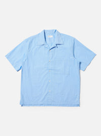 The Pale Blue Broadcloth Camp Shirt is shown laid flat against a plain white background. The short-sleeve shirt features a wide camp collar, a button-down front with a loop closure at the top, and a single large patch pocket on the left chest. The garment has a straight hem with side splits and is made from a smooth, light blue cotton fabric.