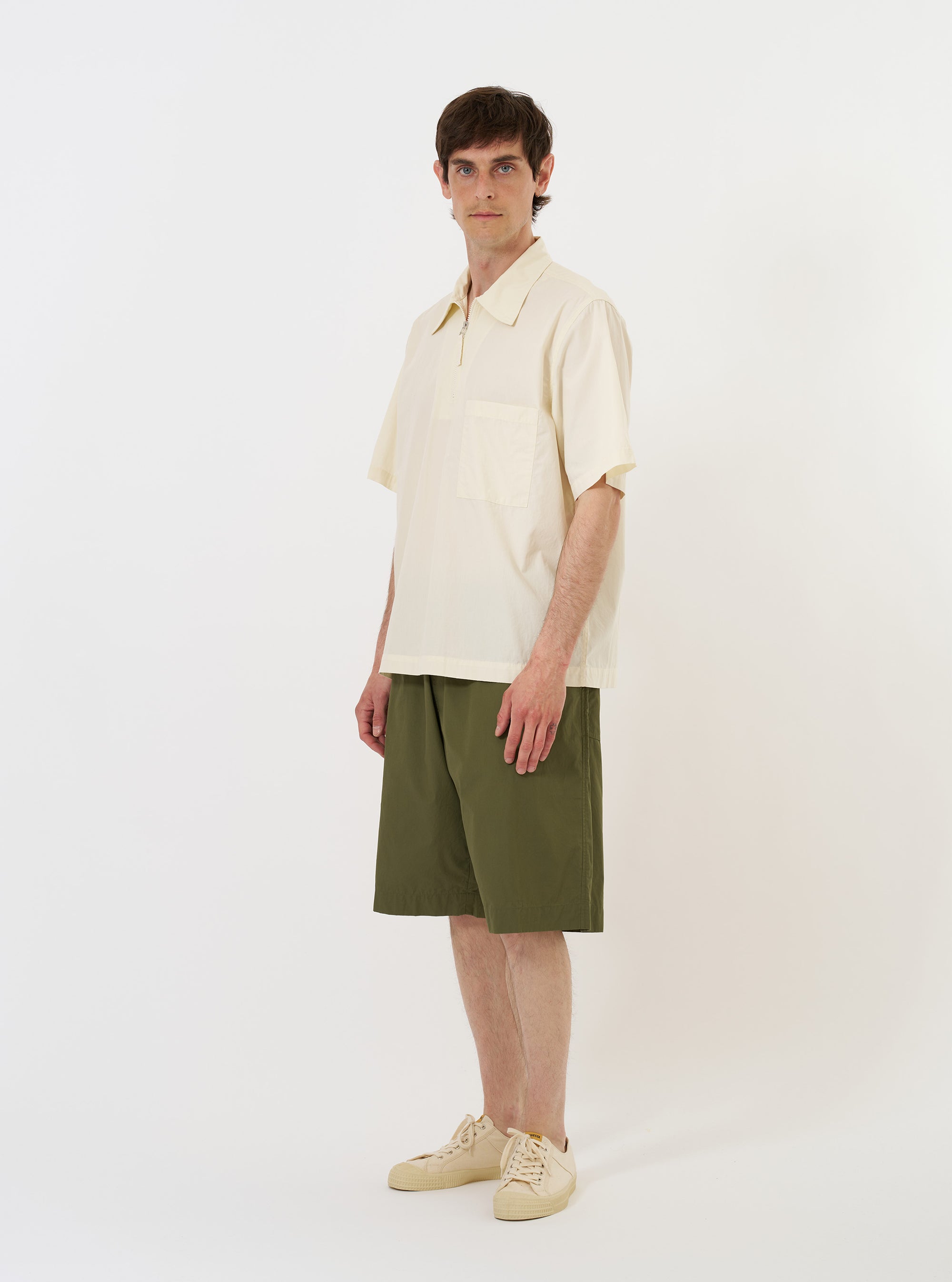 The Ecru Broadcloth Ewan Shirt is shown laid flat against a solid white background. The shirt is a short-sleeved, pullover style featuring a quarter-zip neckline with a silver-toned zipper. It has a classic pointed collar, a single patch pocket on the left chest, and a straight hem with small side splits.