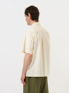 A model is wearing the Ecru Broadcloth Ewan Shirt against a plain white background. The image features the back and side of the garment, highlighting the horizontal back yoke, central box pleat, and short sleeves. The shirt has a boxy, relaxed fit and a straight hem with a side split. The model is also wearing dark-colored trousers.