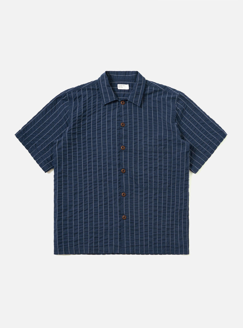 A Navy Chester Stripe S/S Fatigue Shirt is shown laid flat against a solid white background. The garment features a navy blue and white vertical stripe pattern, a classic pointed collar, a full button-down front, and two large chest pockets. The short sleeves are visible, and