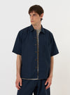 A model is wearing the Navy Paper Touch Cotton S/S Fatigue Shirt against a plain white background. The short-sleeved overshirt is shown from the chest up, featuring a classic pointed collar and two prominent chest pockets with buttoned flaps. The navy blue garment is worn over a white t-shirt, highlighting the crisp texture of the paper touch cotton.