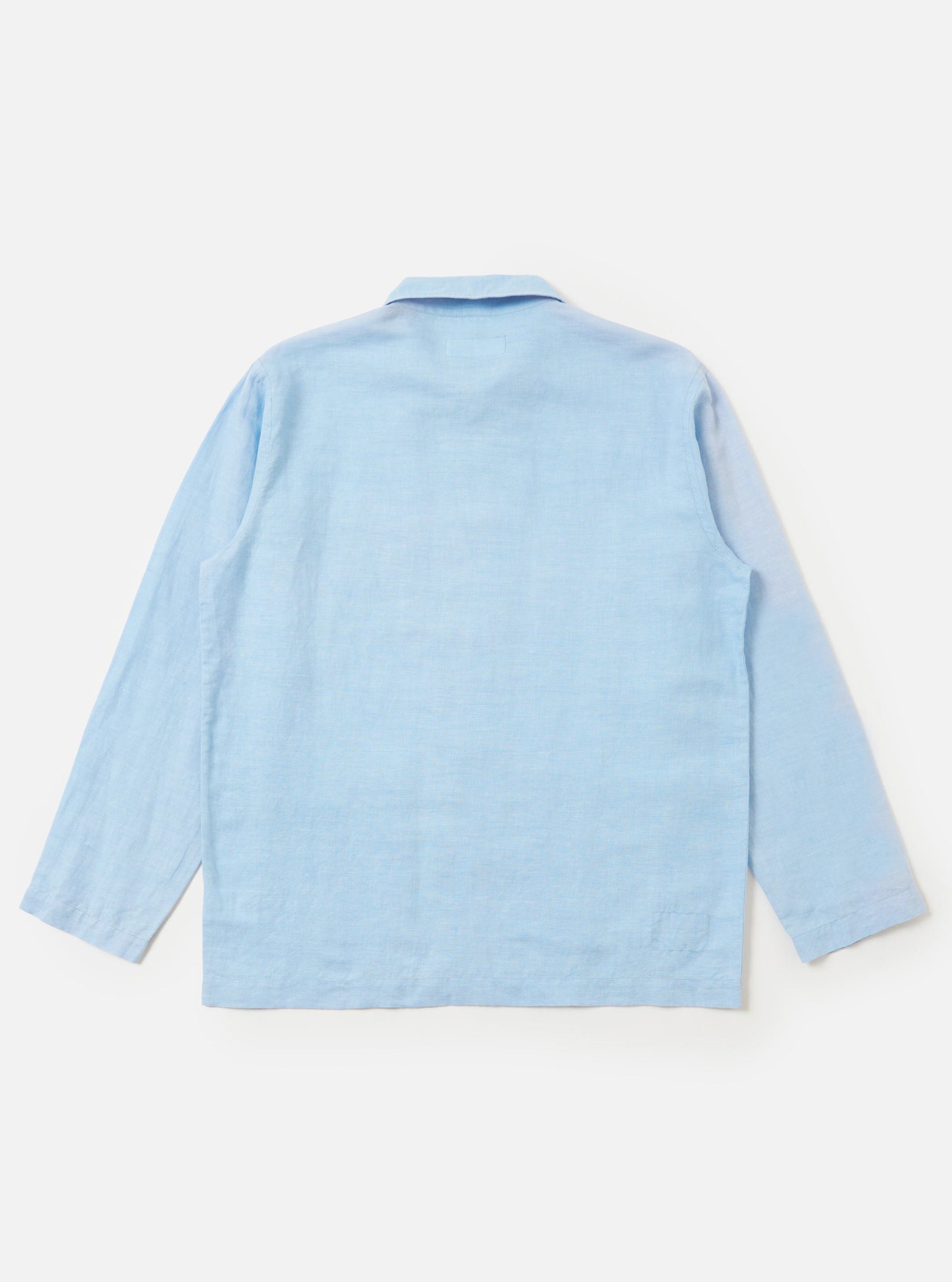 The Pale Blue Linen Cotton Shirting Bakers Overshirt is shown from the back, displayed flat against a plain white background. The image highlights the horizontal back yoke, simple one-piece collar, and the straight hem of the garment. The fabric is a light blue linen-cotton blend.