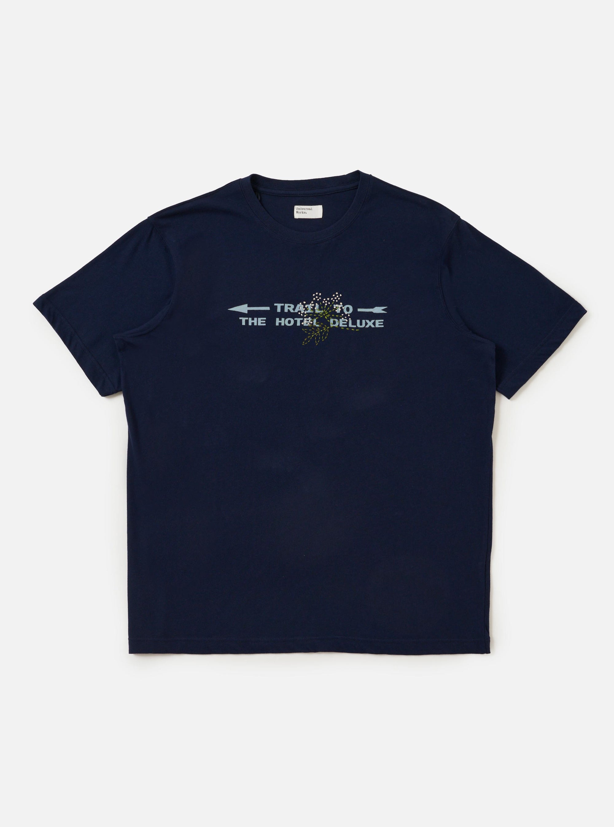 The Navy Single Jersey Trail Trail To Hotel Delux Tee is shown laid flat against a solid white background. The navy blue short-sleeved T-shirt features a classic crew neck and a prominent graphic print on the chest consisting of white text and a circular emblem.