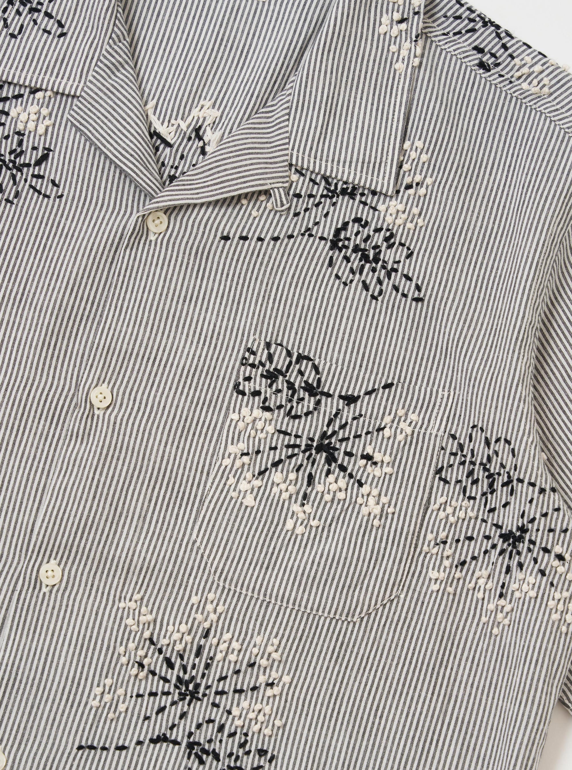 The Grey Hand Embroidered Stripe Cotton Road Shirt is shown from the back, displayed flat against a plain white background. The image highlights the horizontal hand-embroidered stripe pattern in grey and white, the short sleeves, and a straight hem.