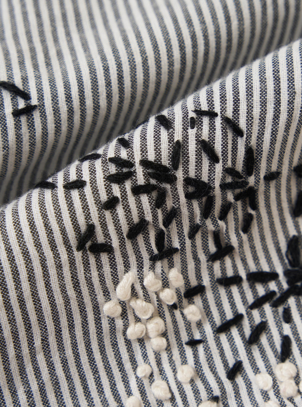 A square fabric swatch of the Grey Hand Embroidered Stripe Cotton Road Shirt shows a detailed close-up of the grey and white vertical striped fabric. Intricate hand-stitched floral embroidery in a darker thread is scattered across the material, highlighting the tactile texture of the cotton.