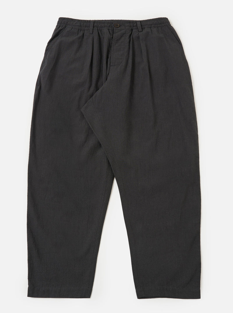 A pair of charcoal grey cotton pleated track pants is displayed flat against a plain white background. The trousers feature an elasticated waistband with a drawstring, a single pleat at the front of each leg, and side pockets.