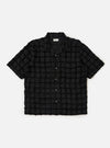 The Black 3D Check Road Shirt is displayed flat against a plain white background. The shirt features a short-sleeve silhouette, a relaxed camp collar, a button-down front, and a single chest pocket. The fabric has a textured 3D check pattern in black and grey.