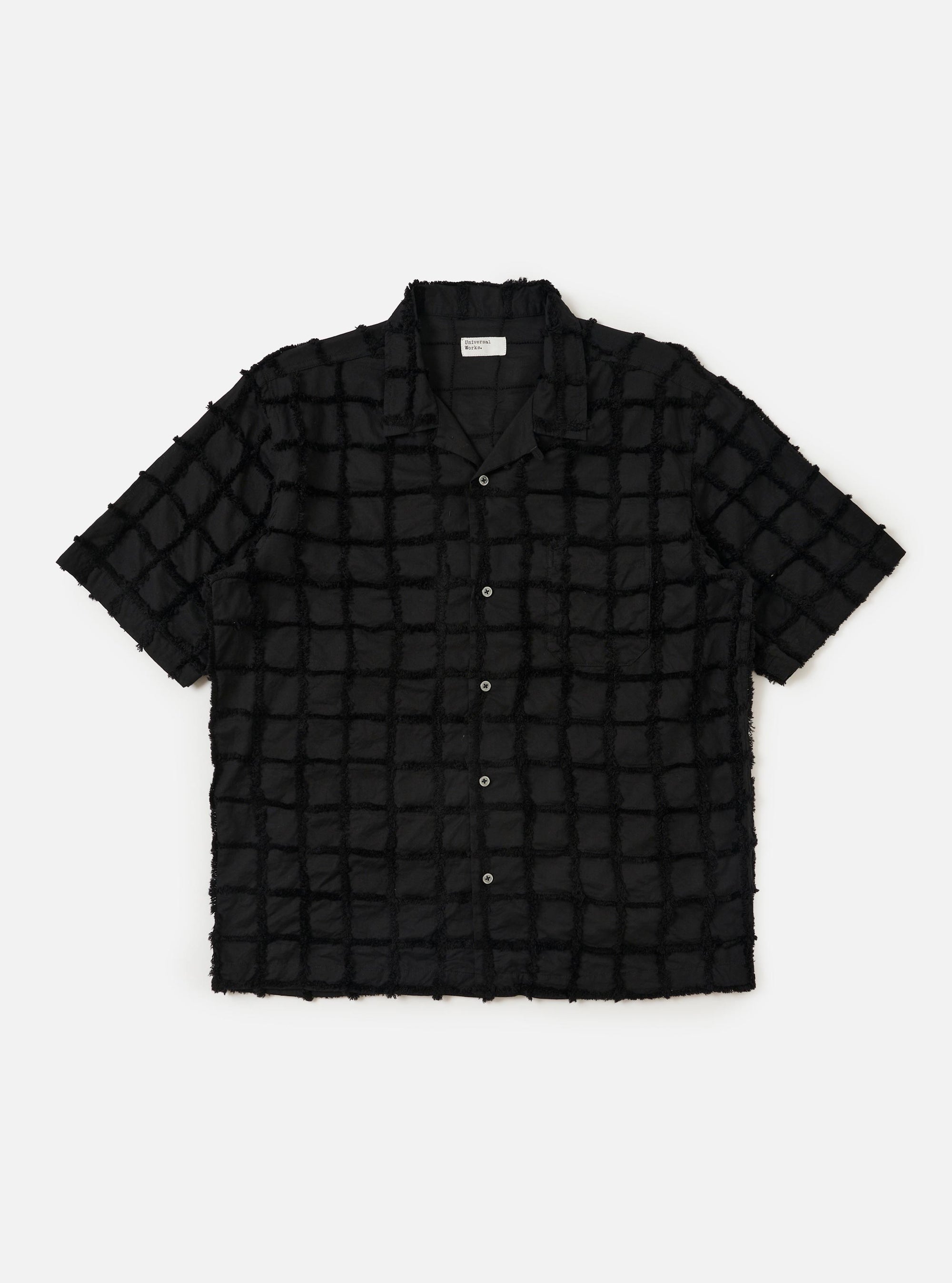 The Black 3D Check Road Shirt is displayed flat against a plain white background. The shirt features a short-sleeve silhouette, a relaxed camp collar, a button-down front, and a single chest pocket. The fabric has a textured 3D check pattern in black and grey.