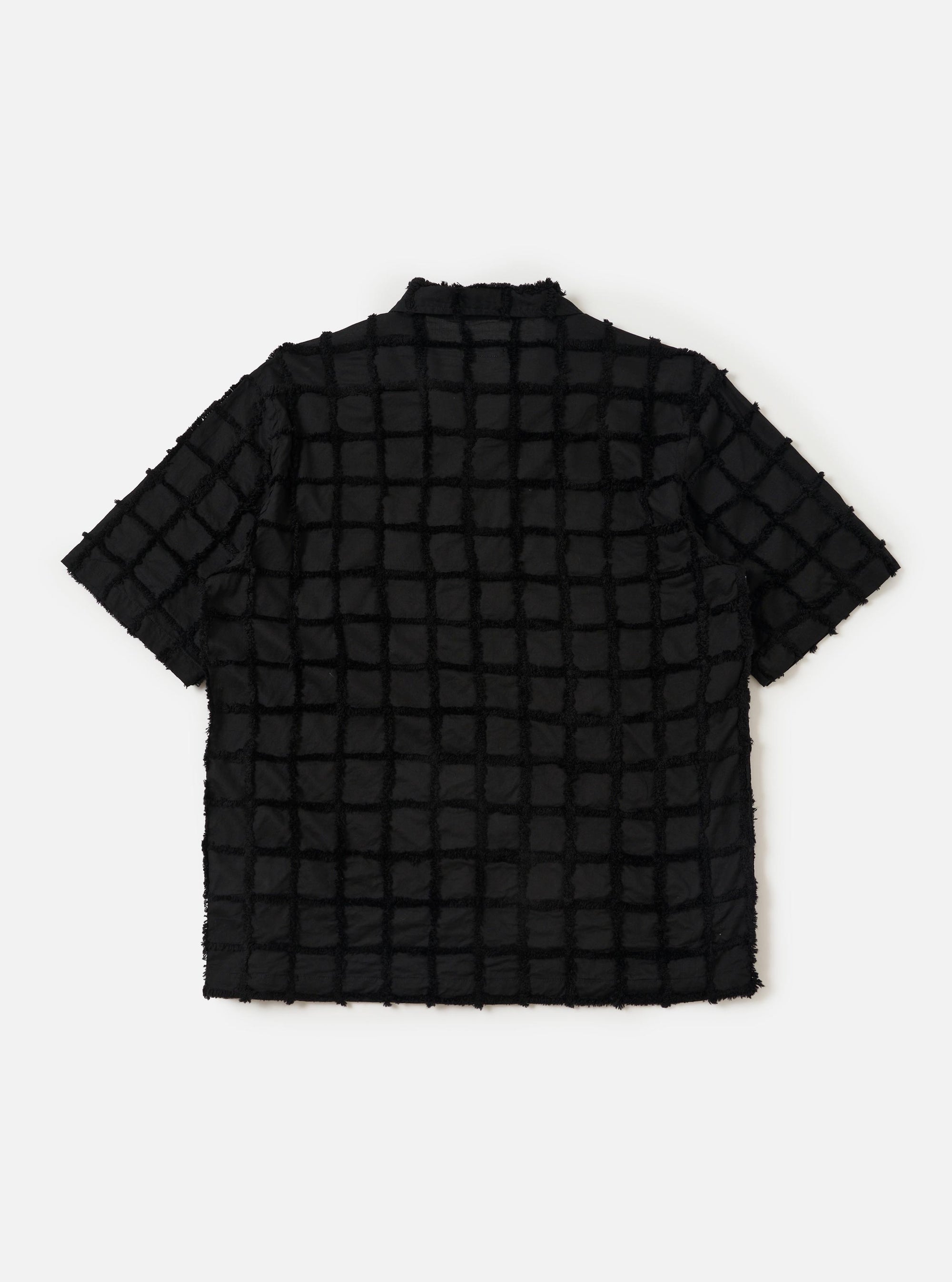 The Black 3D Check Road Shirt is shown from the back, laid flat against a solid white background. The image highlights the shirt's straight hem, short sleeves, and a horizontal yoke across the shoulders. The black fabric features a subtle, textured 3D check pattern throughout.