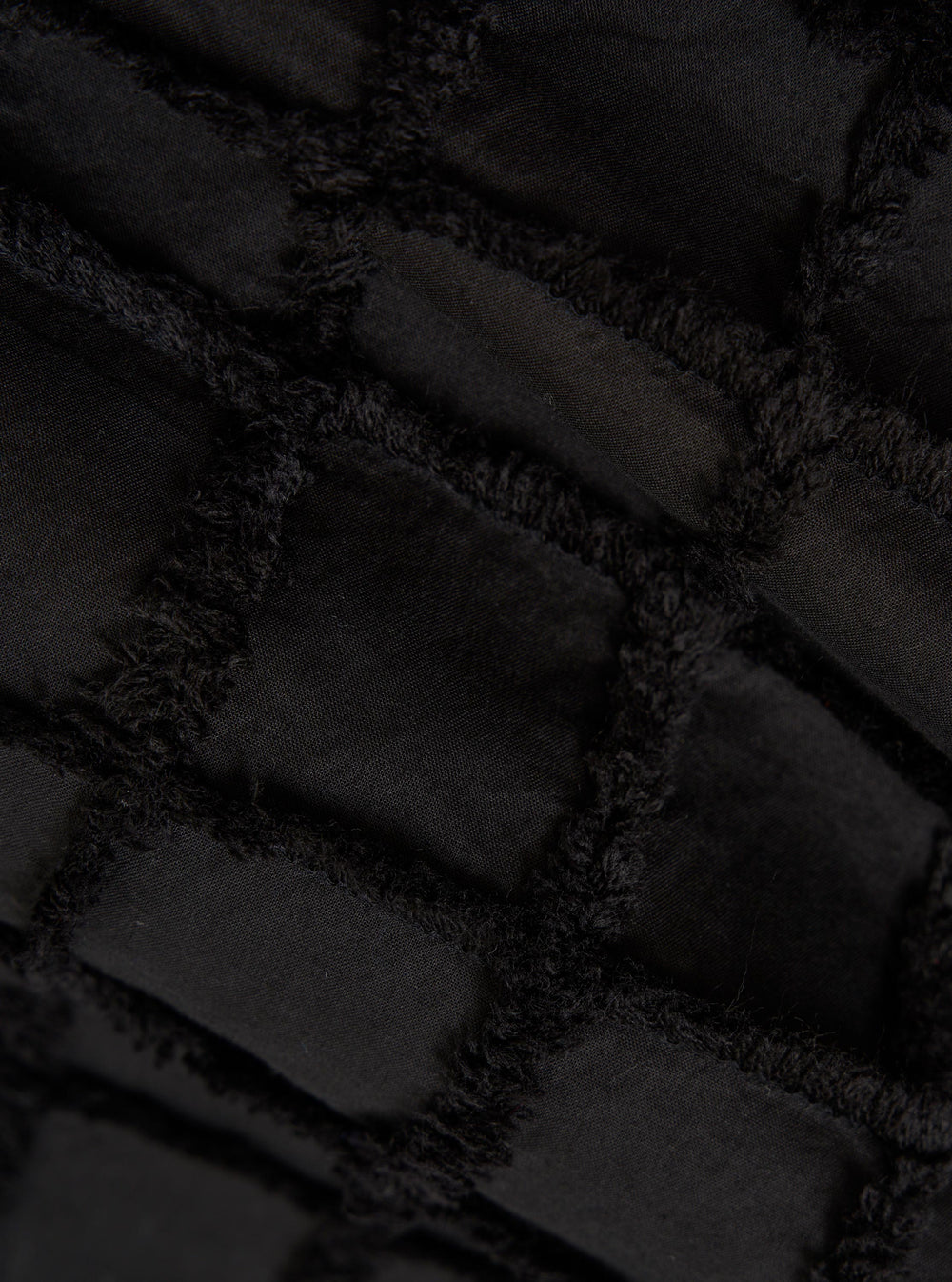 A detailed, close-up fabric swatch of the Black 3D Check Windcheater Jacket highlights a textured cotton material with a three-dimensional grid or checkered pattern in black and dark grey. The image focuses solely on the intricate weave and tonal contrast of the fabric.