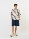 The Pastel Check Patchwork Camp Shirt is shown laid flat against a plain white background. The shirt features a short-sleeve silhouette with a multi-colored patchwork design consisting of various pastel check and plaid patterns in shades of blue, ecru, and pink. Key details include a relaxed camp collar, a full button-down front with a loop-close top button, and a large patch pocket on the left chest. The hem is straight with small side splits.